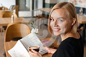 Modern beautiful blond woman reading newspaper while drinking coffee at coffee shop interior, internet distance work