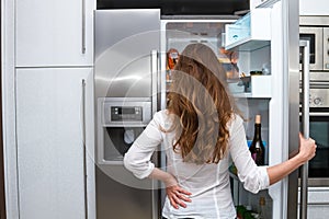 Modern beautiful long haired woman standing and open refrigerators door indoors and looking forward inside the fridge.