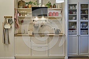 Modern beige kitchen with a living room in a home interior. White cabinets with kitchen utensils and crockery