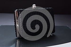 Modern black clutch bag atop a reflective silver surface with a single gold ring in the center