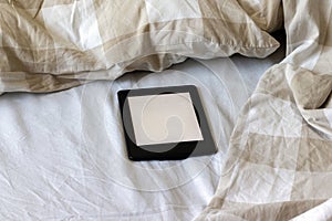 A modern black electronic book with a blank screen on a white and beige bed. Mockup tablet on bedding