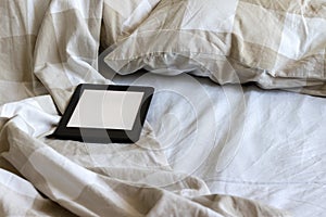 A modern black electronic book with a blank screen on a white and beige bed. Mockup tablet on bedding