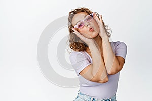 Modern blond girl in sunglasses, pucker lips kissing, making coquettish and flirty face expressions, standing over white