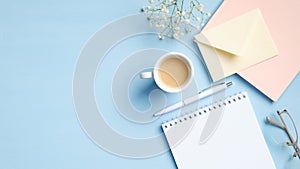 Modern blue office desk with blank notepad, cup of coffee, pink paper notebook, glasses, envelope, flowers. Minimal style feminine