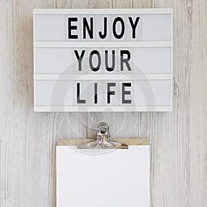 Modern board with text `Enjoy your life`, noticepad with blank sheet of paper on a white wooden background, top view. Overhead,