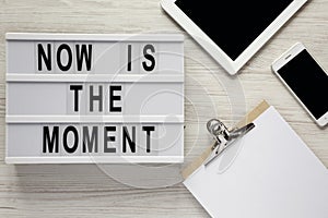 Modern board with text `Now is the moment`, tablet with blank screen, noticepad, smartphone on a white wooden background, top vi