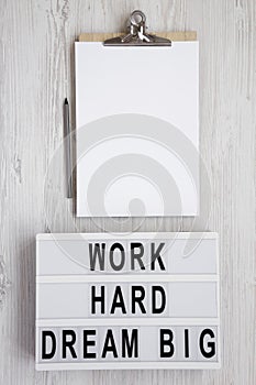 Modern board with `Work hard dream big` words, clipboard with blank sheet of paper on a white wooden background, top view. Flat