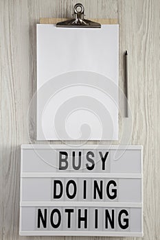 Modern box with text `Busy doing nothing`, noticepad and pencil on a white wooden surface, top view. From above, flat lay,