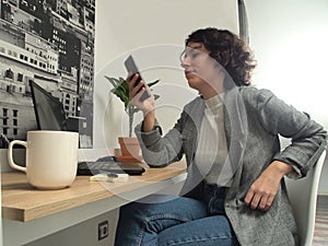 Modern business caucasian woman looking something on her phone with a cup of coffee, post-its and a laptop on her desk