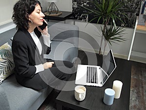 Modern business caucasian woman speaking on the phone, sitting on the sofa with a laptop and a cup of coffee