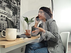 Modern business caucasian woman talking on the phone while is working on her laptop, she has post-its and a cup of coffee on her