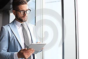 Modern business man in formalwear using digital tablet while standing near window in the office