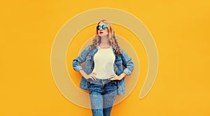 Modern caucasian young blonde woman posing wearing denim jacket, round sunglasses on yellow studio background