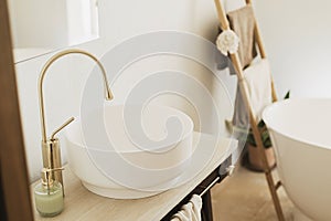 Modern ceramic sink with golden faucet on rustic stand with soap and towel on background of bathtub, modern bathroom interior.