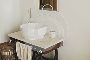 Modern ceramic sink with golden faucet on rustic stand with soap and towel on background of wall with mirror and travertine tile,