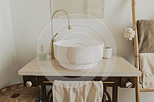 Modern ceramic sink with golden faucet on rustic stand with soap and towel on background of wall with mirror and travertine tile,