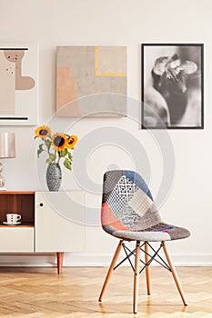 Modern chair in white apartment interior with posters above cupboard with sunflowers. Real photo