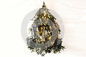 Modern christmas tree made of pine branches with golden festive lights hanging on white wooden wall. Alternative eco christmas