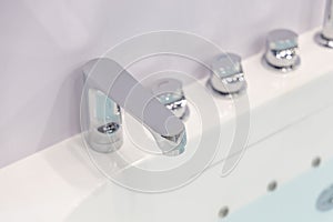 Modern chrome faucet and water taps on a white acrylic bathtub. All parts are made of chromed metal, reflecting the