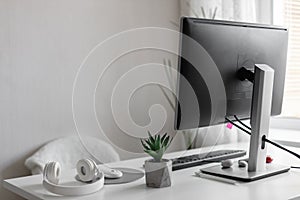 A modern computer monitor is located in a work place in a bright interior. Stylish white wireless headphones and a