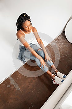 Modern cool American black model woman with stylish dreadlocks in fashion youth casual denim clothes in sneakers sits in room. Top