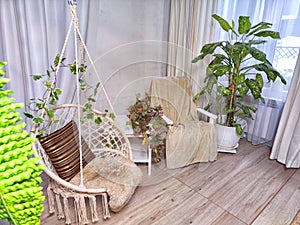 A modern cozy beautiful room with a braided rope macrame swing, chair, green plants, small table and curtains. Interior