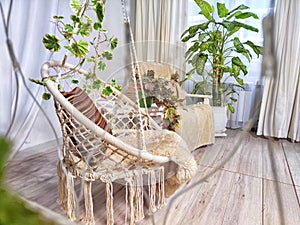A modern cozy beautiful room with a braided rope macrame swing, chair, green plants, small table and curtains. Interior