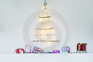 Modern creative christmas eco tree made of wooden sticks hanging on white wall with festive lights and colourful gift boxes.