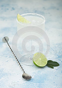 Modern crystal glass of Margarita cocktail with fresh lime and bar spoon on light blue background