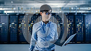 In the Modern Data Center: Portrait of IT Engineer Standing with Server Racks Behind Him, Holding