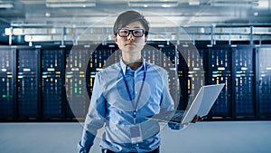 In the Modern Data Center: Portrait of IT Engineer Standing with Server Racks Behind Him, Holding