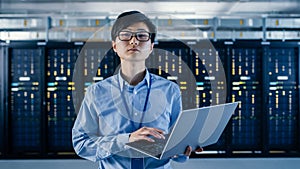 In the Modern Data Center: Portrait of IT Engineer Standing with Server Racks Behind Him, Holding