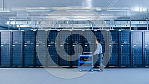 In the Modern Data Center: IT Technician Working with Server Racks, Pushes Cart Between Rows of Se