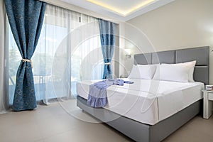 Modern design interior of elegant bedroom with soft grey textile double bed, white bedlinen and blue curtains in classic style