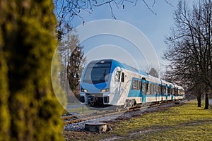 Modern diesel passenger train in white and blue color approaching small station of Rodica between the trees on a sunny winter day