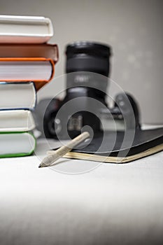 Modern digital SLR camera on the table next to a stack of books, and in the foreground a notebook and pencil.