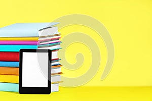 Modern e-book reader and stack of hard cover books on yellow background. Space for text