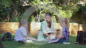 Modern education methods, classmate pupils boy and girl with tutor female read schoolbook and chat during lesson on lawn