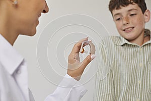 Modern effective hearing aid in hands of female ENT doctor showing it to smiling preteen boy.