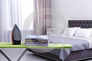 Modern electric iron and clean folded clothes on board in bedroom