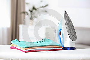 Modern electric iron and clean folded clothes on board in room