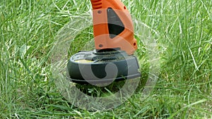 Modern electric orange trimmer with black detail mows green grass. Close-up.