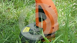 Modern electric orange trimmer with black detail mows green grass. Close-up.