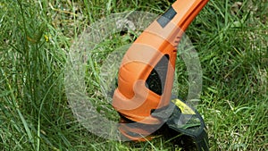 Modern electric orange trimmer with black detail mows green grass. Close-up.