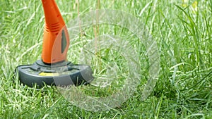 Modern electric orange trimmer with black detail mows green grass. Close-up.