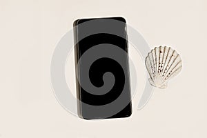 Modern electronic device still life. Black cell phone mock up with sea shell on beige table background in sunlight. Flat