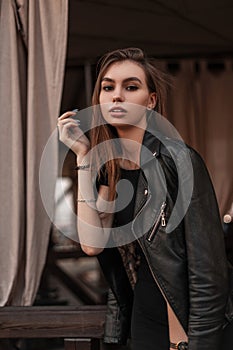 Modern European beautiful young woman in a black fashionable leather jacket in a stylish black dress is resting in a vintage cafe