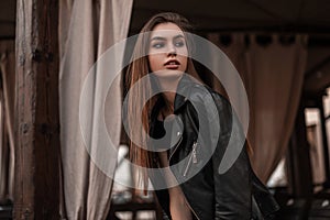 Modern European beautiful young woman in a black fashionable leather jacket in a stylish black dress is resting in a vintage cafe