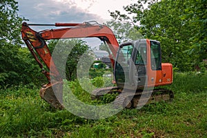 A modern excavator stands on the grass. The bucket is lowered. In the background there are trees and a village house