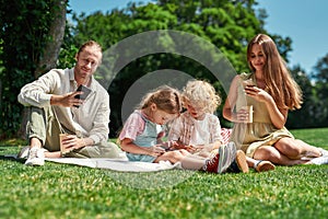 Modern family spending time together outdoors, using mobile devices while having picnic in nature on a summer day